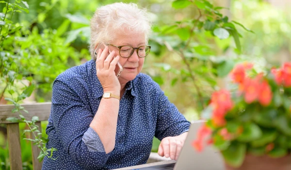 Older Woman In Garden Working On Her Laptop And On The Phone