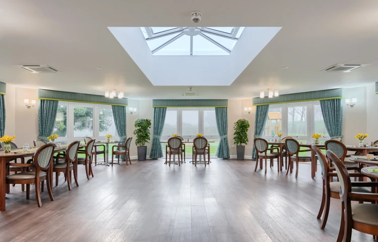 Set tables with chairs in the dining room at Westall House Residential Care Home in Horsted Keynes, featuring a skylight and large windows, where residents enjoy freshly prepared meals