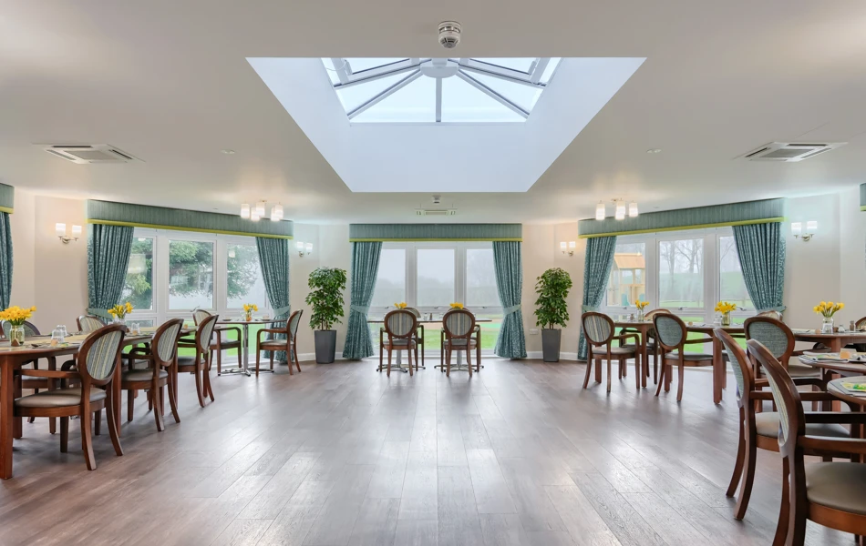 Set tables with chairs in the dining room at Westall House Residential Care Home in Horsted Keynes, featuring a skylight and large windows, where residents enjoy freshly prepared meals